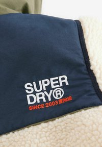 Gros plan sur un tissu bleu marine avec un texte brodé en blanc et orange "SUPER DRY® SINCE 2003" sur un matériau doux en peau de mouton crème.