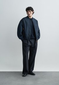 Navy bomber jacket with ribbed cuffs, zip closure, and subtle logo. Paired with dark pleated trousers and black shoes. Smooth fabric texture.