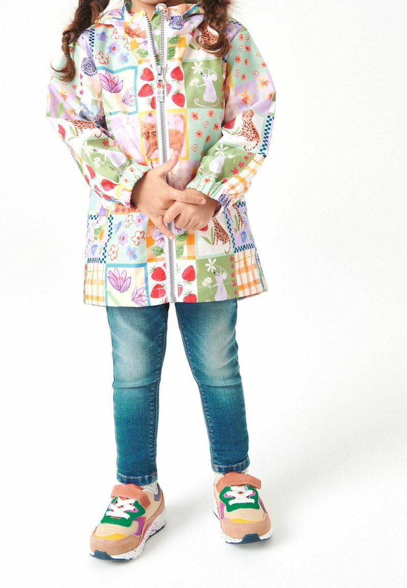 Enfant portant un imperméable patchwork coloré avec des motifs animaux et nature, jean bleu et baskets multicolores, debout les mains jointes.