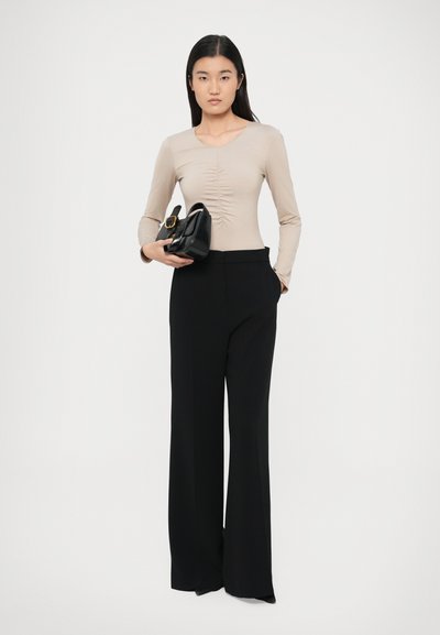 Bézs hosszú ujjú body húzott részletekkel, magas derekú, széles szárú fekete nadrággal párosítva. A modell egy kis fekete kézitáskát tart.