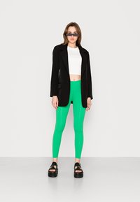 Blazer preto sobre uma blusa cropped branca, combinado com leggings verdes brilhantes e sandálias plataforma pretas. O modelo usa óculos de sol retangulares.