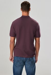 Polo à manches courtes couleur bordeaux, en tissu texturé, avec un col classique et une coupe décontractée ; porté avec un jean bleu.