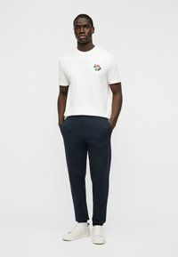 Man stands with hands in pockets, wearing white graphic t-shirt, dark navy pants, and white sneakers against plain background.