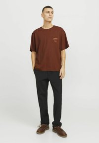 Camiseta marrón de corte holgado y mangas cortas, con un pequeño logo impreso. Lleva puesta con pantalones negros y zapatos marrones.