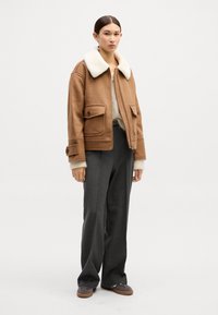 Bruine wollen jas met witte bontkraag, ritssluiting, twee voorkkeeping; gecombineerd met grijze hoge taille broek enbruine schoenen.