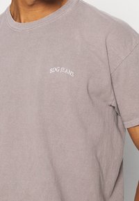 Tričko s krátkým rukávem a klasickým výstřihem v tlumené levandulové barvě z bavlny, se malým bílým logem "BDG JEANS" na hrudi. Měkký, uvolněný střih.