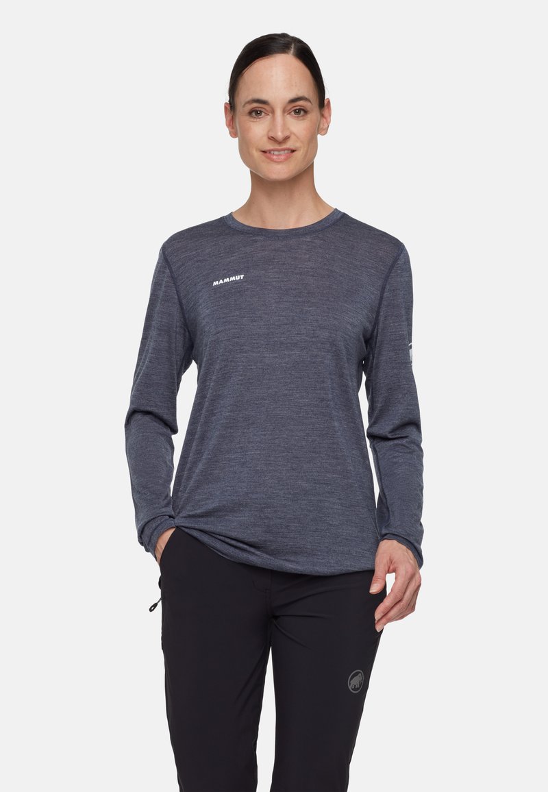 Kobieta ubrana w ciemnoszarą koszulkę Mammut z długim rękawem i czarne spodnie, stojąca z jedną ręką w kieszeni, uśmiechająca się do aparatu.