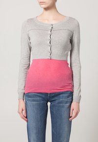 Grå kort cardigan med knappstängning över en rosa, åtsittande topp. Bärs med blå jeans, som visar en jämn textur och enkel design.