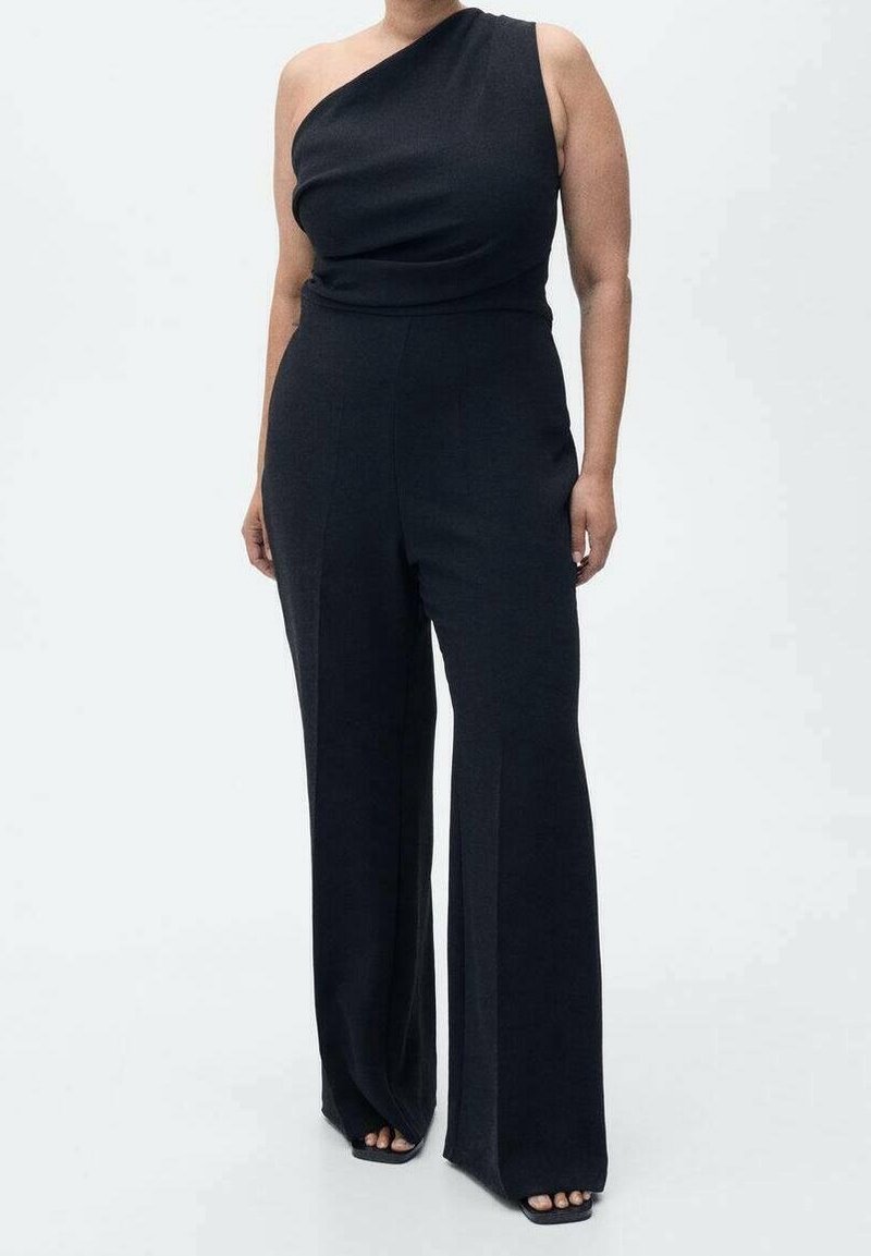 Personne portant une combinaison noire à une épaule avec un pantalon à jambes larges et des chaussures noires ouvertes, se tenant devant un fond clair uni.