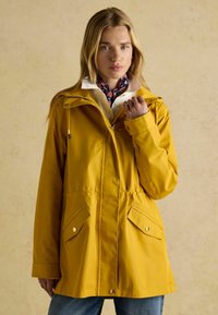 Geltona vandeniui atspari striukė su kapišonu, turinti surištą liemenį, dvi priekines kišenes ir spaustukus. Dėvima ant balto marškinėlio su apykakle.