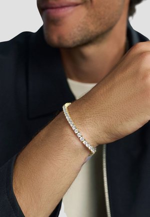 Man wearing a gold tennis bracelet with large clear gemstones on wrist, dressed in dark jacket and light shirt.