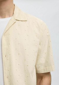 Camicia a maniche corte in morbido tessuto beige chiaro con un motivo testurizzato di fiori in rilievo e piccole aperture, dotata di colletto classico e pattina con bottoni.