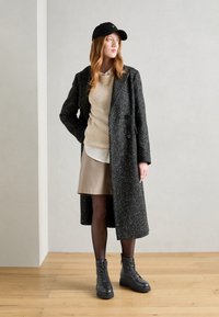 Manteau long en mélange de laine noir avec des taches blanches, superposé à un pull beige à côtes et une jupe beige. Des bottes noires à lacets complètent le look.