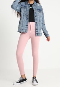 Chaqueta de mezclilla azul claro con detalles desgastados, camiseta negra, jeans ajustados rosas y zapatillas bajas negras.