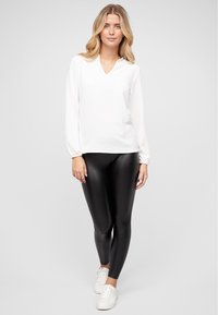 Maglietta bianca a maniche lunghe con scollatura a V, abbinata a leggings neri lucidi e sneakers bianche. La maglietta ha una vestibilità comoda e maniche arricciate.