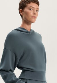 Felpa con cappuccio in morbido tessuto teal tenue. Presenta una vestibilità comoda, spalle scese e una superficie liscia. Nessun motivo o accentuazione visibili.