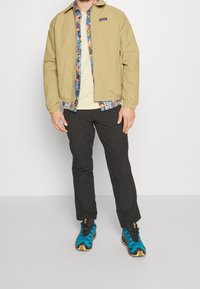 Tan zip-up jacket over a patterned shirt, paired with black pants and blue athletic shoes. Jacket features elastic cuffs and a collar.