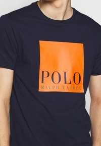 Navyn blå t-shirt med en orange ruta som visar "POLO RALPH LAUREN" bärs av en person som visas från halsen till midjan.