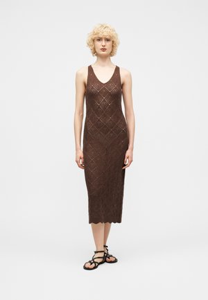 Donna con capelli biondi ricci che indossa un vestito midi marrone senza maniche in maglia e sandali neri bassi con cinturini, in piedi davanti a uno sfondo bianco semplice.