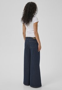Woman with long curly hair wearing a white t-shirt and wide-leg dark blue jeans, standing facing away on a plain background.
