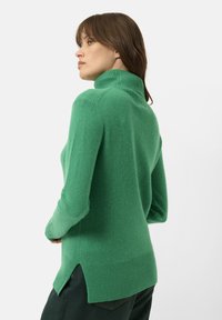 Maglione verde a collo alto realizzato in materiale morbido, con orlo a costine e spacco laterale per facilitare i movimenti, mostrato da un angolo laterale.