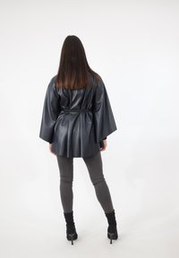 Schwarze Jacke aus Kunstleder mit Kimono-Schnitt, tailliertem Bund und weiten Ärmeln, kombiniert mit grauer, enger Hose und schwarzen Stiefeletten.