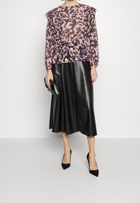 Lila und khaki abstrakt gemusterte, durchsichtige Bluse mit Rüschensäumen, kombiniert mit einem schwarzen, fließenden Leder-Midirock und schwarzen Absätzen.