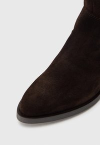 Botín de ante marrón con punta puntiaguda, que presenta una suela fina de goma y una textura suave. El detalle de costuras a lo largo del cuello realza el diseño.