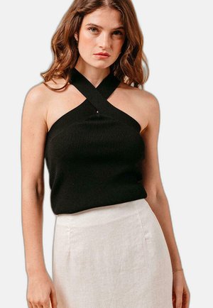 Vrouw draagt een zwarte mouwloze haltertop gecombineerd met een lichtbeige rok met hoge taille, poserend tegen een effen achtergrond.