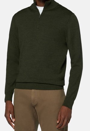 Man draagt een olijfgroen trui met kwart rits over een wit overhemd en lichtbruine broek, staat met ontspannen armen langs het lichaam.