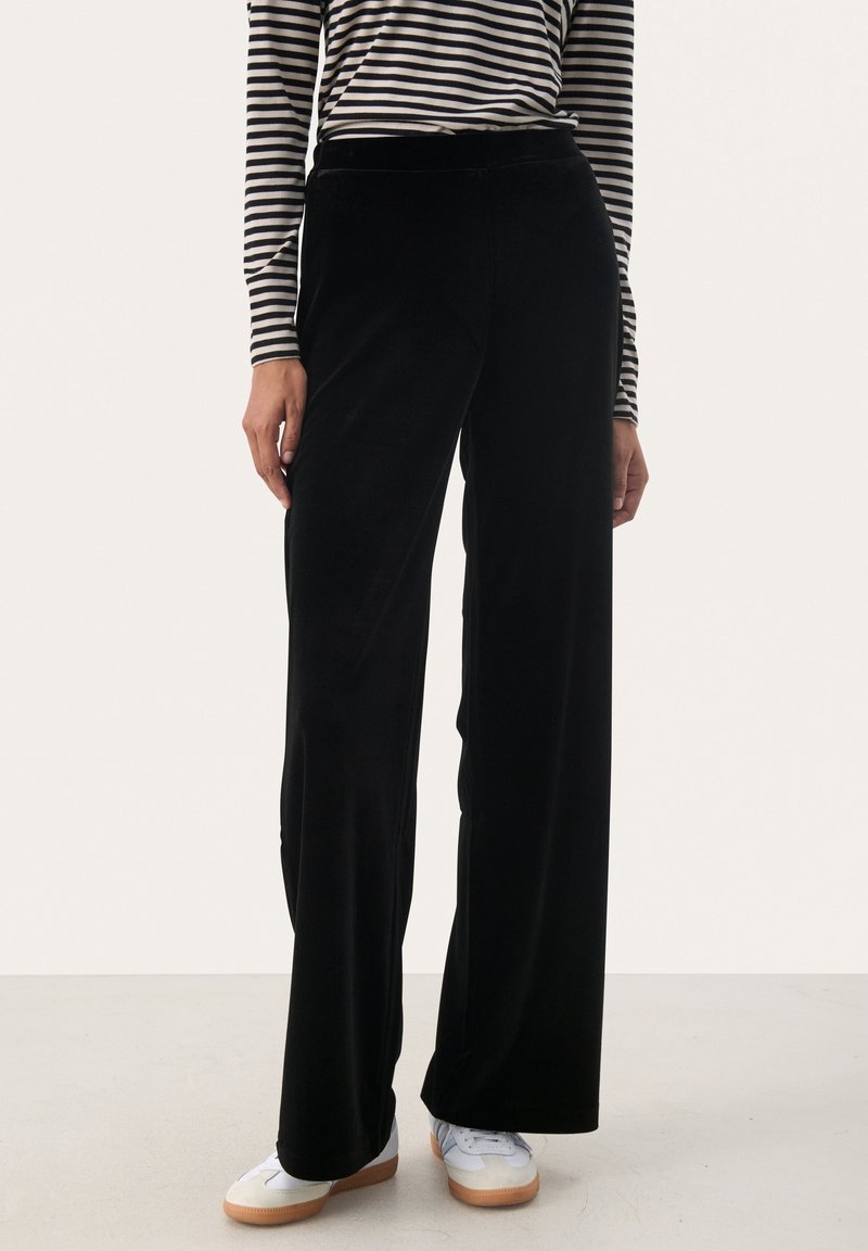 Femme portant un pantalon large noir, des baskets blanches et un haut à manches longues rayé noir et blanc, debout sur un sol clair.