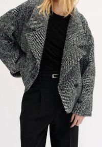 Svartvit houndstooth oversize blazer med breda slag, dubbelknappning och texturerad tyg, kombinerad med en svart topp och byxor.
