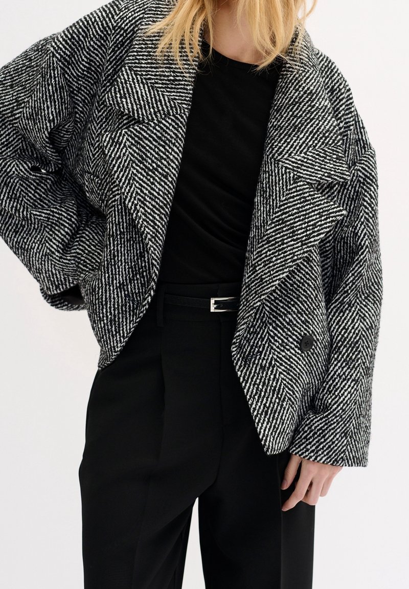 Svartvit houndstooth oversize blazer med breda slag, dubbelknappning och texturerad tyg, kombinerad med en svart topp och byxor.