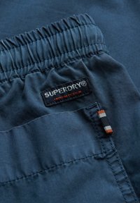 Tissu bleu avec une taille froncée arborant une étiquette indiquant "SUPERDRY" et un accent avec des rayures orange et noires. Texture douce.