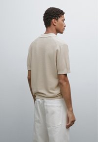 Beige stickad polo skjorta med korta ärmar, med en ribbad krage och ett strukturerat mönster. Bärs med vita byxor, visad bakifrån.