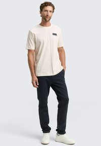 Beige t-shirt met korte mouwen en een rechthoekige blauwe grafiek op de borst, gecombineerd met donkerblauwe broek en witte sneakers.