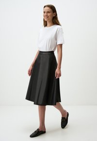 Jupe A-line en faux cuir noir avec une texture lisse, de longueur genou, associée à un t-shirt blanc et des chaussures noires à enfiler.