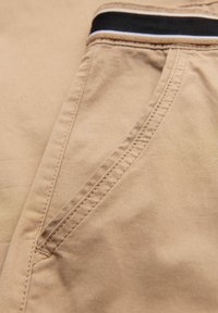 Close-up af beige bukselomme og søm med sort-hvid stribet linningdetalje på stoffet.