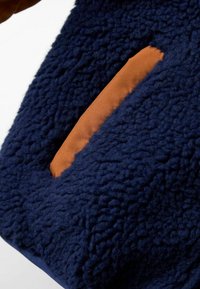 Jūras spēku teksturēts audums ar pūkainu izskatu, ar brūnu mākslīgās ādas kabatu. Kontrastējošie materiāli izceļ dizainu.