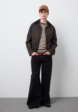 Jeune personne portant une casquette marron, une veste sombre boutonnée, une chemise rayée et un pantalon large noir, debout les mains dans les poches devant un fond uni.