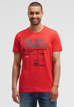 Smiling man with short hair wearing a red Camp David t-shirt with text and icons, standing against a plain light background.