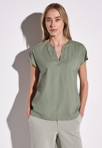 Top corto verde chiaro a maniche corte con scollatura a V e tessuto morbido. La modella lo indossa con pantaloni grigi chiari e a vestibilità rilassata.