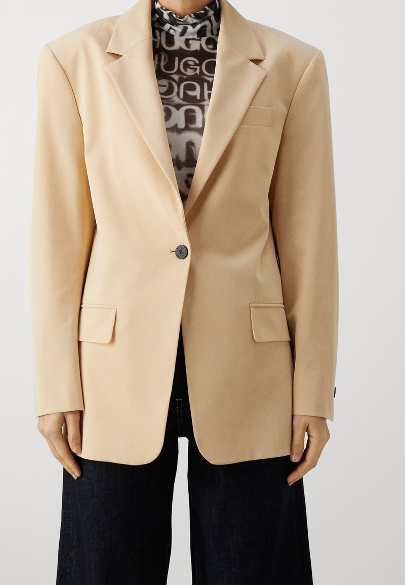 Femme portant un blazer beige à un bouton sur un col roulé à motifs noir et blanc et un pantalon large foncé, debout devant un fond uni.