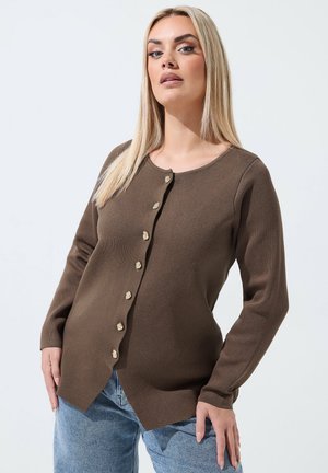 Kvinde med langt blond hår iført brun langærmet knappet cardigan og lyseblå jeans står mod en lys baggrund.