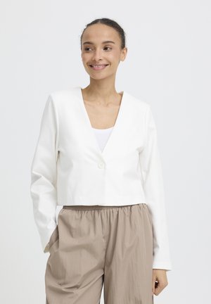 Naeratav naine, kes kannab valget lühikest bleiserit valge topi ja lahtiste beežide pükstega, seisab ühevärvilise hele tausta ees.