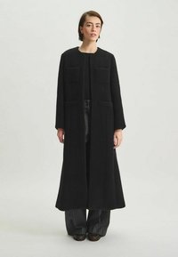 Manteau noir long en tissu texturé, avec deux poches avant, une coupe droite et un style ouvert, associé à un pantalon noir.