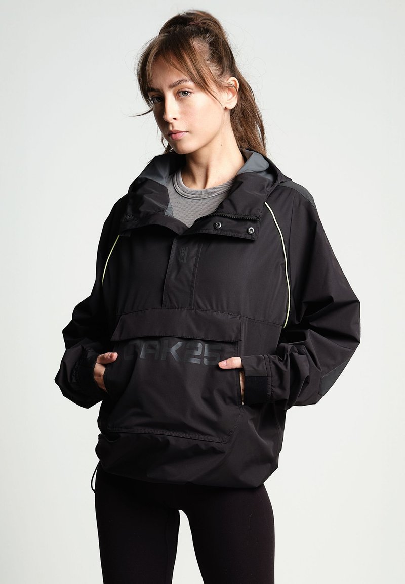 Mujer joven con el cabello castaño recogido, vistiendo una chaqueta negra de gran tamaño con un bolsillo frontal y leggings negros, de pie contra un fondo liso.