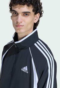 Melnas sportiskas jakas ar baltām svītrām, augsti stāvošu apkakli un Adidas logo uz krūtīm. Gluda tekstūra un eleganta silueta.