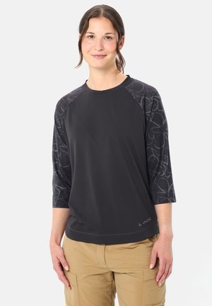 Femme portant un chemisier noir à manches trois-quarts avec des épaules à motifs et un pantalon cargo beige, debout devant un fond uni.