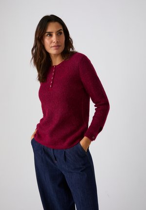 Femme portant un pull en maille rouge avec des boutons et un pantalon bleu, debout les mains dans les poches, regardant sur le côté sur un fond uni.
